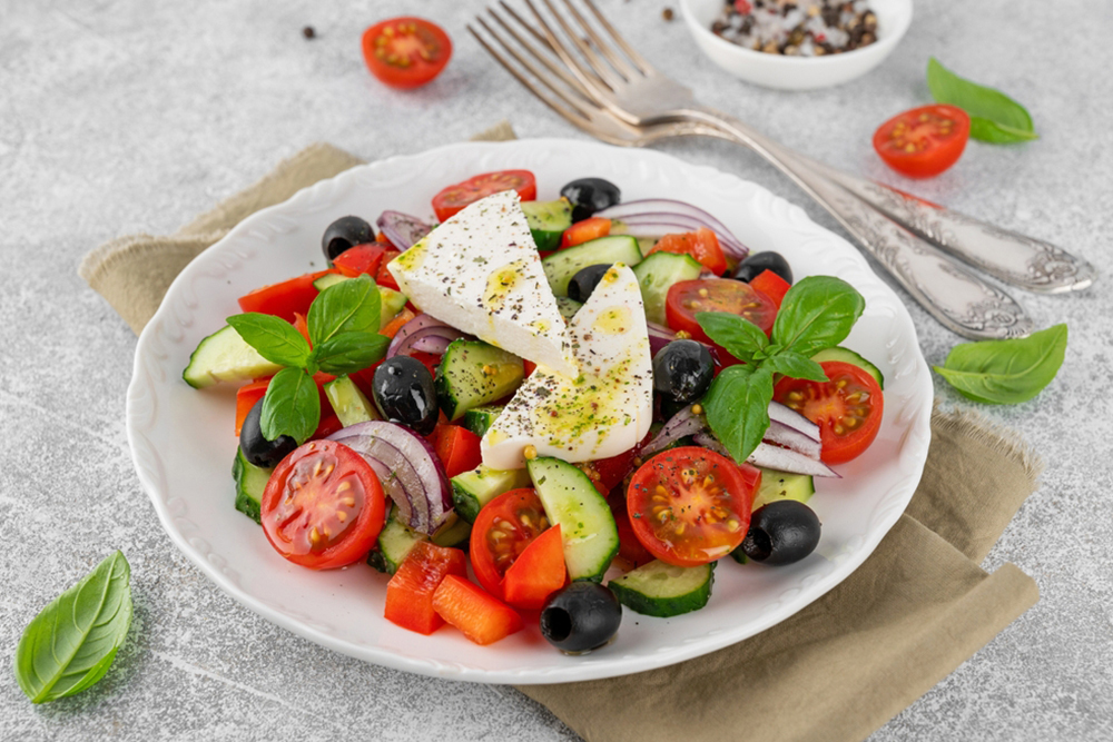 Greek salad of fresh juicy vegetables, feta cheese, herbs and olives on a white plate on a light concrete background. Healthy food. Copy space, top view